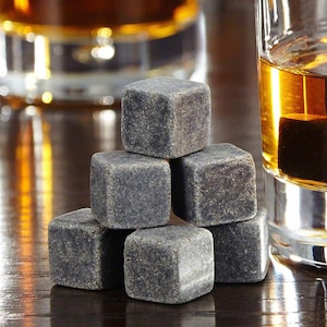 May include: A set of six gray stone whiskey chilling cubes. The cubes are stacked in a pyramid shape on a wooden surface. A glass of amber liquid is in the background.
