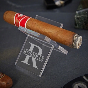 May include: A clear acrylic cigar holder with a personalized engraved initial "R" and the name "ROBERTS" below. A brown cigar is resting on the holder.
