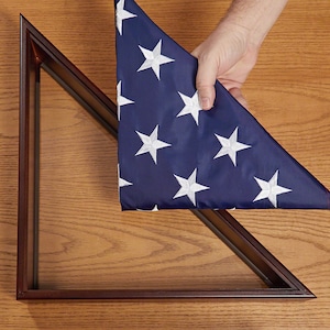 May include: A dark wood triangular display case with a folded American flag. The flag is blue with white stars and red and white stripes. The case is empty, ready to hold the flag.