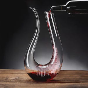May include: A clear glass wine decanter with a long, curved spout is being filled with red wine from a bottle. The decanter has a wide base and a slender neck. The initials "RDA" are etched on the front of the decanter. The background is dark.
