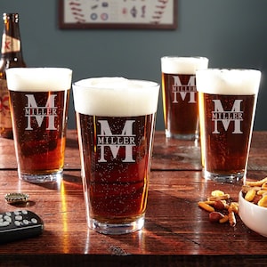 May include: Four clear glass beer mugs with a personalized monogram and the name "Miller" etched on the front. The mugs are filled with a dark amber beer and are sitting on a wooden table.