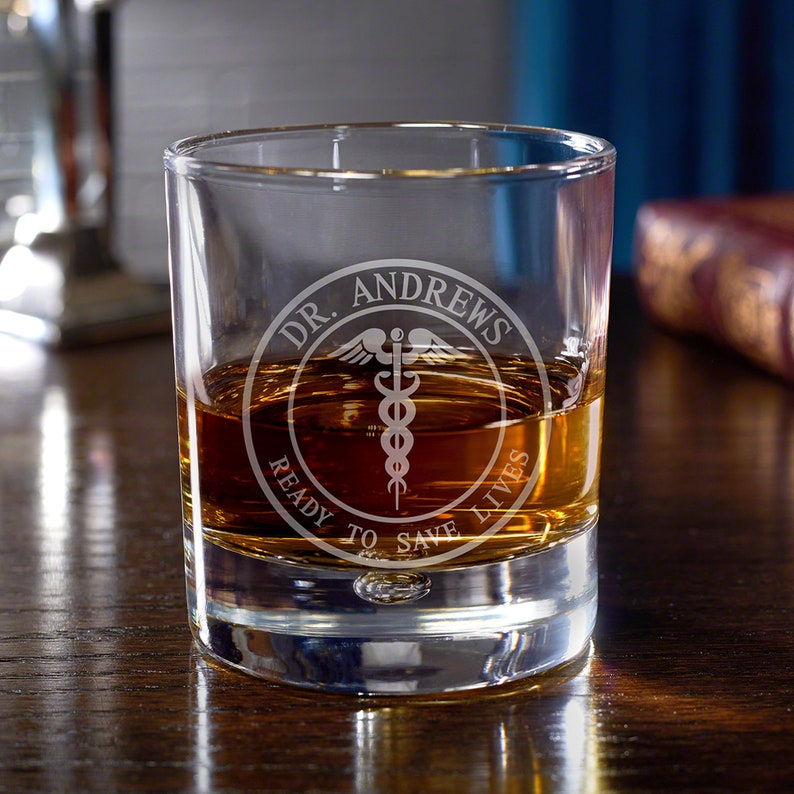 May include: A clear glass rocks glass with a personalized inscription that reads "Dr. Andrews Ready to Save Lives" with a medical symbol in the center.