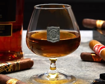 May include: A clear glass brandy snifter with a silver initial "G" on the side, filled with amber liquid. The snifter is sitting on a dark surface with cigars in the background.