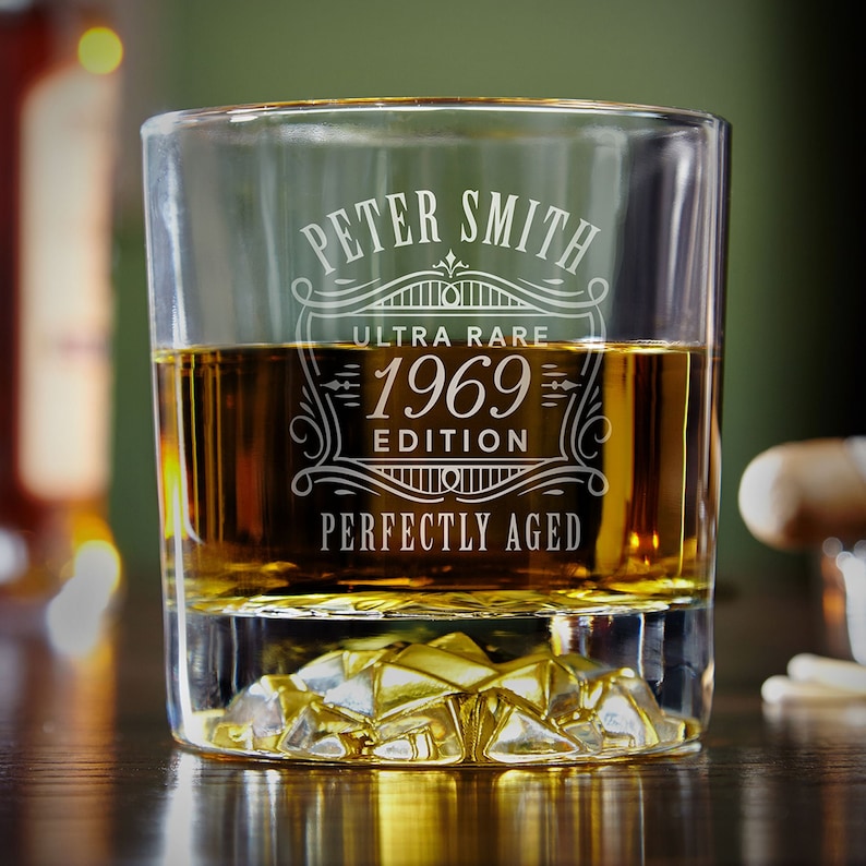 May include: A clear glass rocks glass with a personalized inscription that reads "Peter Smith Ultra Rare 1969 Edition Perfectly Aged". The glass is filled with amber-colored liquid.