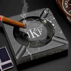 May include: A black marble ashtray with a round depression in the center and three smaller depressions around the edge. The ashtray is engraved with the initials "JKF". A lit cigar is resting on the edge of the ashtray.