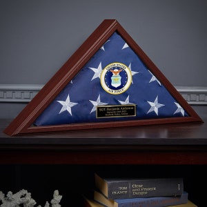May include: A wooden triangular shadow box with a folded American flag inside. The flag is blue with white stars and has the United States Air Force emblem in the center. A gold plaque with the text "SGT. Benjamin Anderson September 24th 2021 Husband, Father, Airman" is attached to the bottom of the box.