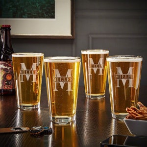 May include: Four clear glass beer mugs with a personalized etched design. Each mug features the letter "M" and the word "Miller" etched into the glass.
