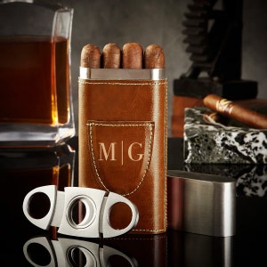 May include: A brown leather cigar case with a silver metal top and a silver metal cigar cutter. The case has a monogram "M|G" on the front.