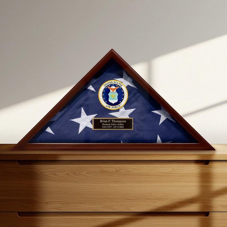 May include: A triangular wooden flag display case with a United States Air Force emblem and a folded American flag. The case includes a plaque with the name Brian F. Thompson and dates 9/21/1977-12/17/2021.