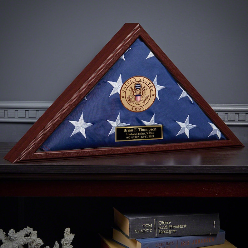 May include: A wooden triangular flag display case with a folded American flag inside. The flag has white stars on a blue field. A gold-colored plaque with the text "UNITED STATES ARMY" and a nameplate with text are visible.