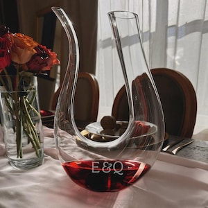 May include: A clear glass wine decanter with a curved design, filled with red wine. The base of the decanter has the initials "E&Q" etched into the glass. A vase of flowers is visible in the background.