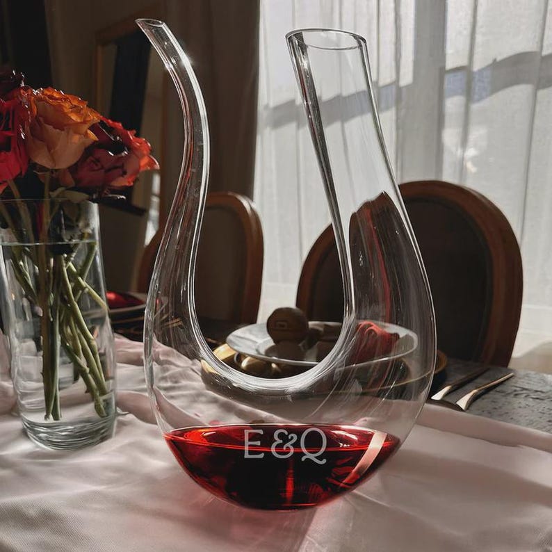 May include: A clear glass wine decanter with a curved design, filled with red wine. The base of the decanter has the initials "E&Q" etched into the glass. A vase of flowers is visible in the background.