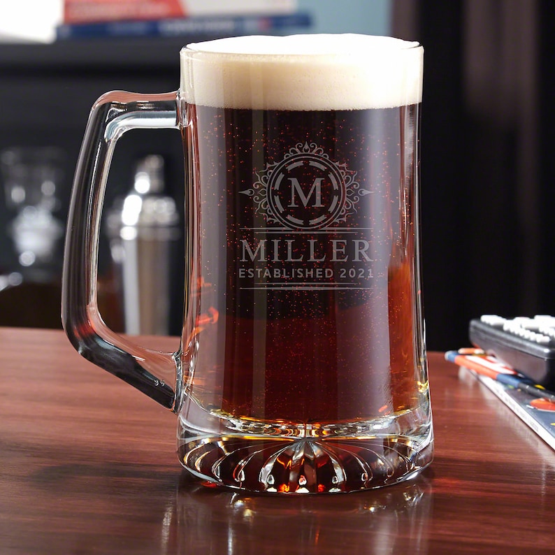May include: A clear glass beer stein with a handle. The stein is filled with a dark brown beer and has a white head. The stein is engraved with a monogram and the text "Miller Established 2021."