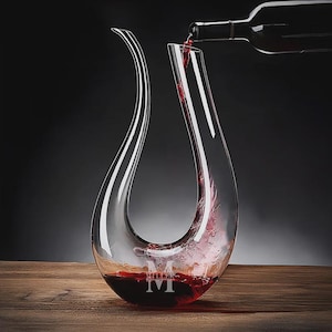 May include: A clear glass wine decanter with a long, curved spout is being filled with red wine from a dark bottle. The decanter has a wide base and a monogrammed "M" at the bottom. The background is dark, and the decanter sits on a wooden surface.