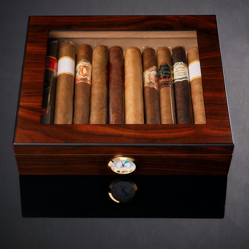 May include: A wooden humidor with a glass top, revealing a collection of brown cigars. The humidor has a built-in hygrometer to monitor humidity levels.
