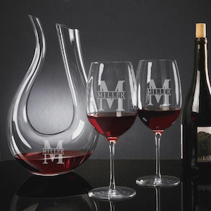 May include: A clear glass wine decanter and two wine glasses filled with red wine. The glasses and decanter are engraved with the letter "M" and the word "MILLER". A bottle of wine with a cork is also visible.