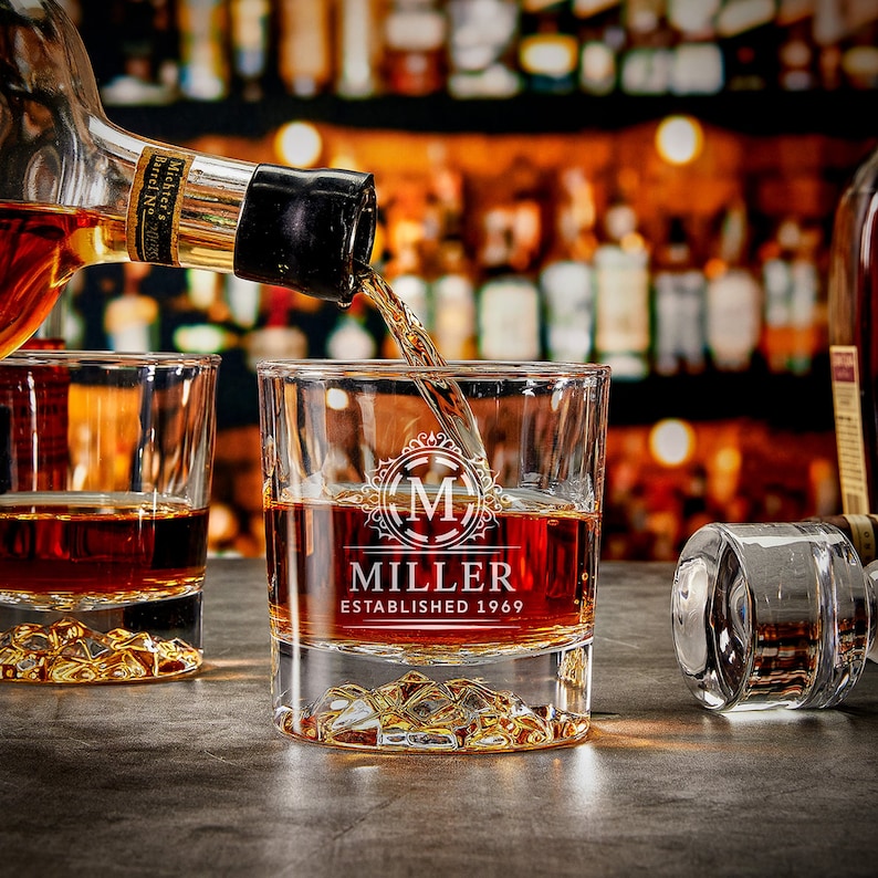 May include: A clear glass whiskey tumbler with a monogrammed design. The glass has the text "MILLER ESTABLISHED 1969" etched on the front. The glass is filled with amber liquid and ice cubes.
