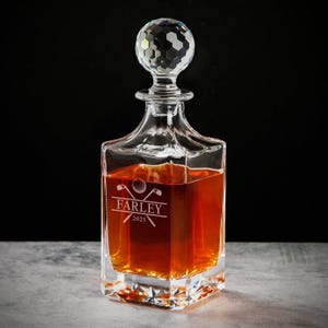 May include: A clear glass decanter with a faceted crystal stopper. The decanter is filled with amber liquid and features the name "FARLEY" and the year "2025" with golf-themed graphics. The decanter sits on a gray surface.