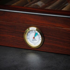 May include: A gold-colored humidor hygrometer with a white face and black markings. The needle is pointing to 65%. The text on the face reads "Humidor Hygrometer" and "rel. %"