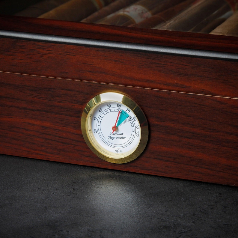 May include: A gold-colored humidor hygrometer with a white face and black markings. The needle is pointing to 65%. The text on the face reads "Humidor Hygrometer" and "rel. %"