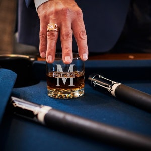 May include: A personalized whiskey glass with the name "MATTHEWS" and the initial "M" etched on the front. The glass is filled with amber liquid and is sitting on a blue surface.