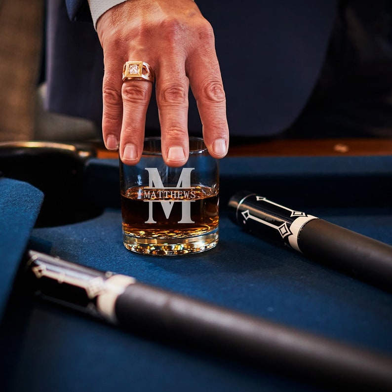 May include: A personalized whiskey glass with the name "MATTHEWS" and the initial "M" etched on the front. The glass is filled with amber liquid and is sitting on a blue surface.