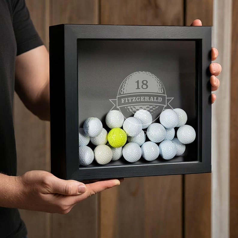 May include: A black shadow box with a glass front filled with golf balls. The box has a personalized inscription with the number "18" and the name "FITZGERALD".