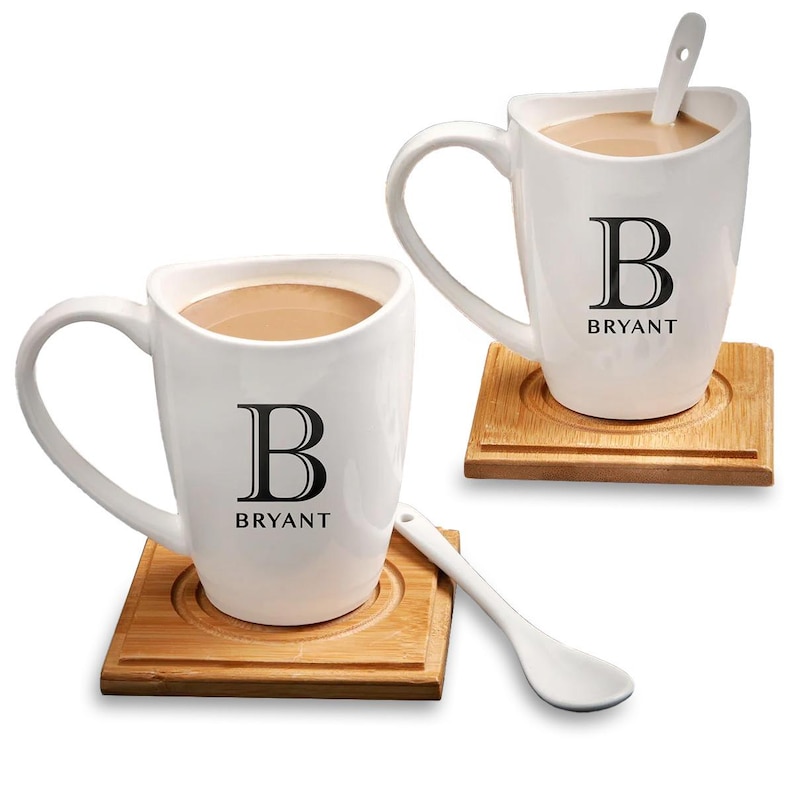 K&ouml;nnte beinhalten: Zwei wei&szlig;e Keramikbecher mit schwarzen Griffen und einem schwarzen Buchstaben "B" sowie dem Wort "BRYANT" aufgedruckt auf der Vorderseite. Jeder Becher steht auf einem Untersetzer aus Holz und enth&auml;lt einen wei&szlig;en Keramikl&ouml;ffel.