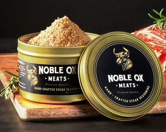 May include: A gold-coloured tin of "NOBLE OX MEATS" hand-crafted steak seasoning, with a black label and a bull logo. The lid is off, revealing a pile of brown seasoning. A wooden chopping board and a piece of raw steak are in the background.