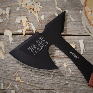 May include: Black axe with a wooden handle. The axe has a black blade with the text "Max Smith Best Man 11.9.2023" engraved on it. The axe is on a wooden surface with wood shavings.