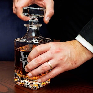 May include: A clear glass decanter with a square base and a stopper. The decanter is filled with amber liquid and has the name "WILLI" etched on the front.