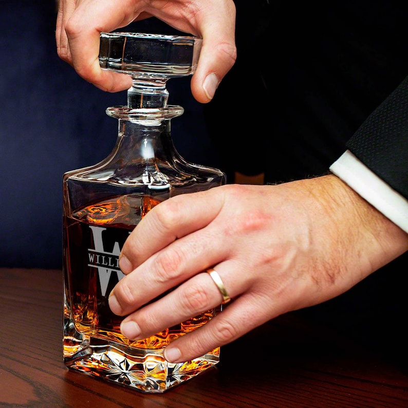 May include: A clear glass decanter with a square base and a stopper. The decanter is filled with amber liquid and has the name "WILLI" etched on the front.