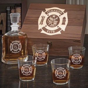 May include: A wooden box with a firefighter emblem engraved on the lid. The box contains a glass decanter and four glasses, all with the same firefighter emblem.