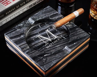 May include: Black marble ashtray with a gold trim and a personalized inscription that reads "M MATTHEWS". A cigar is resting on the ashtray.