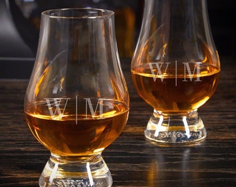 May include: Two clear, tulip-shaped whiskey glasses filled with amber liquid. Each glass has the initials "WM" etched on the front. The glasses sit on a dark wooden surface, suggesting a bar or home setting. The base of one glass reads "GLENCAIRN".