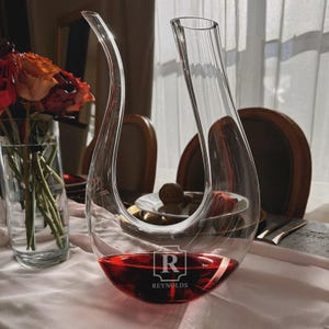 May include: A clear glass wine decanter with a curved spout and a wide base, filled with red wine. The decanter has the word "REYNOLDS" and the letter "R" etched on the base. A vase of flowers is visible in the background.