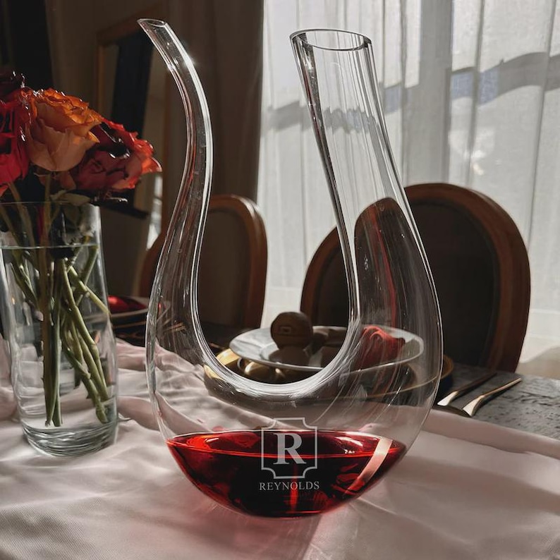 May include: A clear glass wine decanter with a curved spout and a wide base, filled with red wine. The decanter has the word "REYNOLDS" and the letter "R" etched on the base. A vase of flowers is visible in the background.
