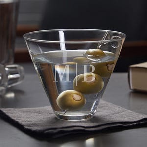 Może przedstawiać: Szklanka do martini z przezroczystego szkła z srebrnym szpikulcem do oliwek i zielonymi oliwkami. Szklanka jest grawerowana inicjałami "M|B".
