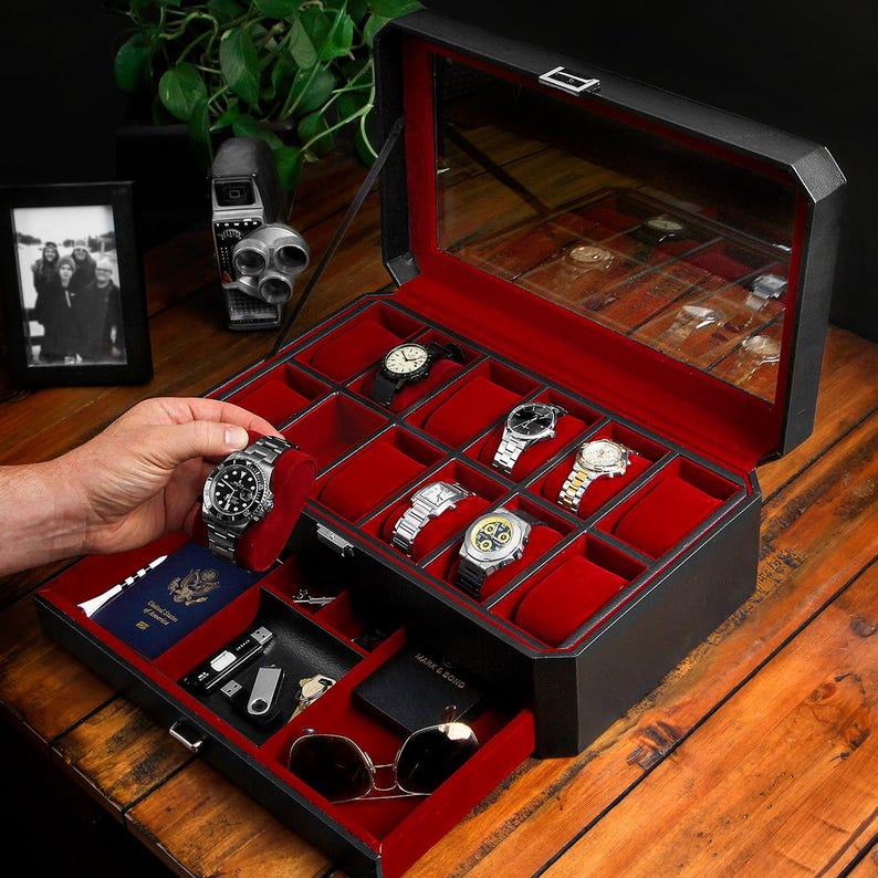 Puede incluir: Caja de reloj de cuero negro con interior de terciopelo rojo y tapa de cristal. La caja tiene un caj&oacute;n con forro de terciopelo rojo y un peque&ntilde;o compartimento para gafas de sol. La caja est&aacute; abierta y contiene varios relojes.