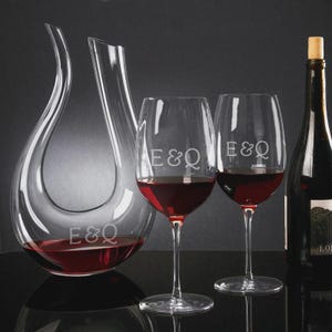 May include: A clear glass wine decanter and two wine glasses filled with red wine. The decanter has a unique curved design. The glasses and decanter are etched with the letters "E&Q". A bottle of wine with a cork is also present.