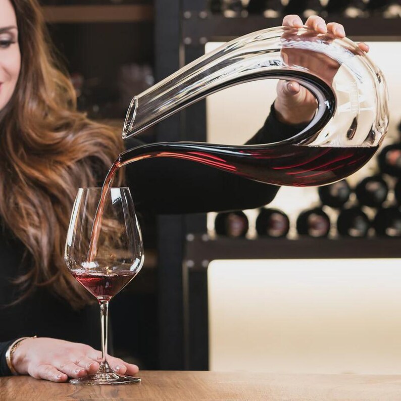 May include: A clear glass wine decanter pouring red wine into a stemmed wine glass. The decanter has a curved design and is held by a person. The wine glass is on a wooden surface.