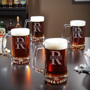 May include: Four clear glass beer mugs with handles. Each mug is filled with a dark amber beer and has a white foam head. The mugs are etched with the letter "R" and the word "ROBERTS".
