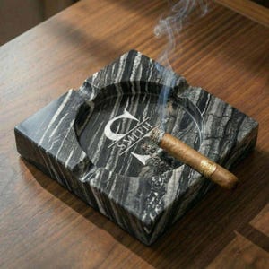 May include: A square, black and gray marble ashtray with a lit cigar resting inside. The ashtray is engraved with the letter "C" and the name "SMITH". Smoke rises from the cigar, and the ashtray sits on a wooden surface.