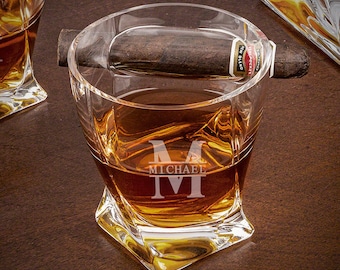 May include: A clear glass whiskey tumbler with a personalized engraved initial "M" and the name "MICHAEL". The glass is filled with amber liquid and a cigar is resting on the rim.