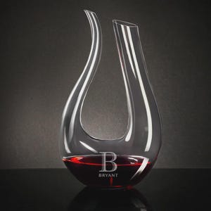 May include: A clear glass wine decanter with a unique, curved design. The decanter is filled with red wine and features the name "BRYANT" and the letter "B" etched on the front. The decanter is on a black surface against a dark gray background.