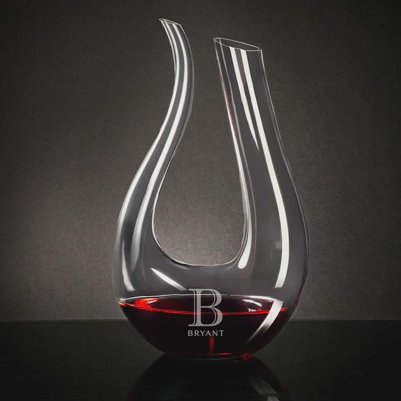 May include: A clear glass wine decanter with a unique, curved design. The decanter is filled with red wine and features the name "BRYANT" and the letter "B" etched on the front. The decanter is on a black surface against a dark gray background.