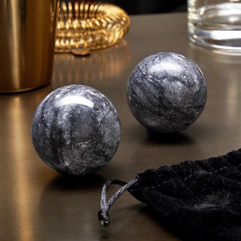May include: Two gray marble massage balls with black veining, resting on a dark surface next to a black velvet pouch.