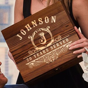 Pu&ograve; includere: Una scatola di legno con un'iscrizione personalizzata. L'iscrizione recita "JOHNSON 20 YEARS SERVED" con una iniziale decorativa "J" al centro. La scatola ha una finitura in legno a grana marrone scuro.
