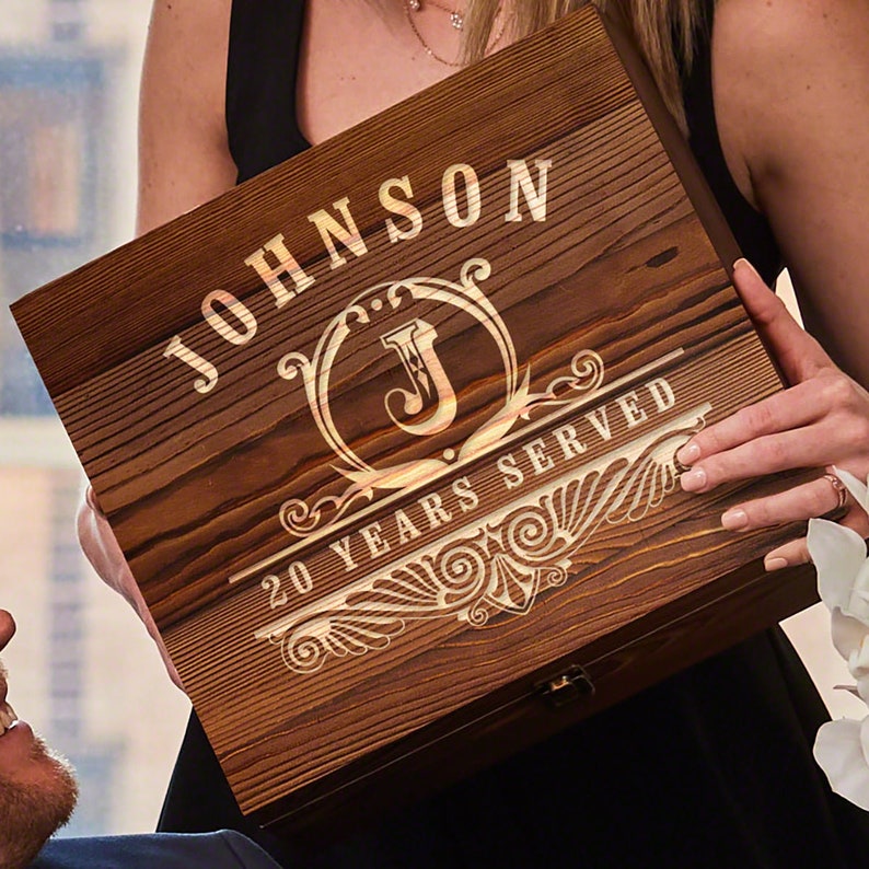 Pu&ograve; includere: Una scatola di legno con un'iscrizione personalizzata. L'iscrizione recita "JOHNSON 20 YEARS SERVED" con una iniziale decorativa "J" al centro. La scatola ha una finitura in legno a grana marrone scuro.