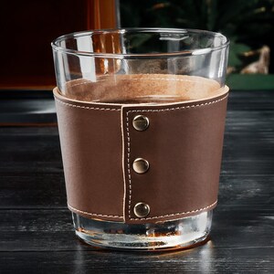 May include: A clear glass tumbler with a brown leather wrap and three brass rivets. The leather wrap has a stitched seam.